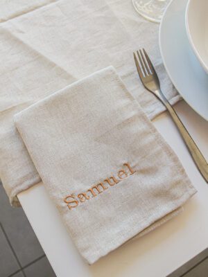 Serviette Mathieu plié et déposé à côté d'une assiette blanche avec une broderie en marron : Samuel.